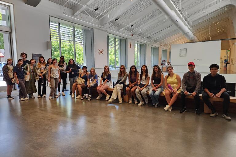 Group photo of high school students who participated in our Berkeley Changemaker Pre-College program. Half of the students are standing, half of the students are sitting on a long bench in the hallway.